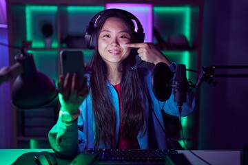 Young asian woman playing video games with smartphone pointing with hand finger to face and nose, smiling cheerful. beauty concept