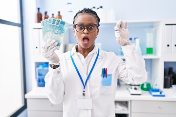 African woman with braids working at scientist laboratory holding money in shock face, looking skeptical and sarcastic, surprised with open mouth