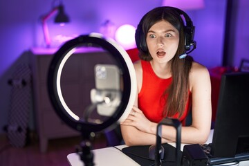 Young caucasian woman playing video games recording with smartphone scared and amazed with open mouth for surprise, disbelief face