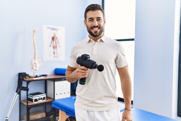 Naklejka na ściany i meble Handsome hispanic man holding therapy massage gun at physiotherapy center looking positive and happy standing and smiling with a confident smile showing teeth