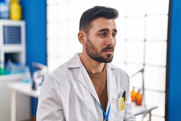 Young hispanic man wearing scientist uniform with serious expression at laboratory