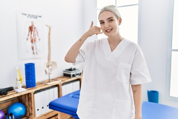 Young caucasian woman working at pain recovery clinic smiling doing phone gesture with hand and fingers like talking on the telephone. communicating concepts.