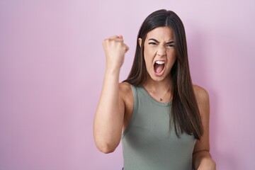 Hispanic woman standing over pink background angry and mad raising fist frustrated and furious...