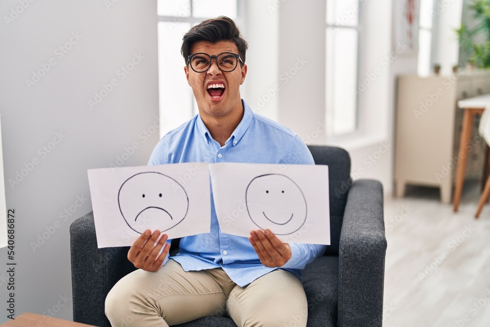 Wall mural Hispanic man working on depression holding sad to happy emotion paper angry and mad screaming frustrated and furious, shouting with anger looking up.