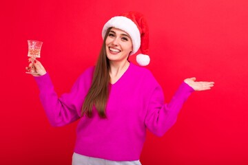 A young happy woman in a pink sweater in a Santa Claus hat holds a glass of champagne on a red background