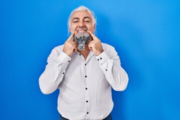 Middle age man with grey hair standing over blue background smiling with open mouth, fingers pointing and forcing cheerful smile