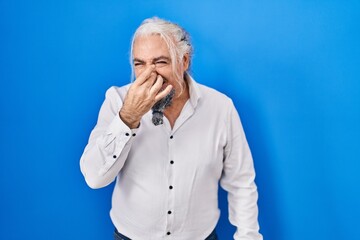 Middle age man with grey hair standing over blue background smelling something stinky and disgusting, intolerable smell, holding breath with fingers on nose. bad smell