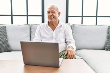 Senior man using laptop at home sitting on the sofa winking looking at the camera with sexy...