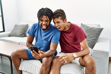 Young hispanic men using smartphone sitting on the sofa at home angry and mad screaming frustrated...