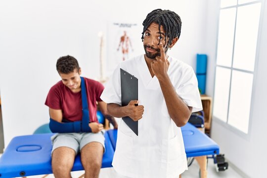 Young hispanic man working at pain recovery clinic with a man with broken arm pointing to the eye watching you gesture, suspicious expression