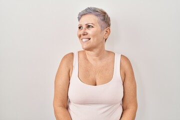 Middle age caucasian woman standing over white background looking away to side with smile on face, natural expression. laughing confident.