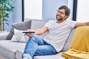 Handsome latin man holding television remote control smiling with a happy and cool smile on face....