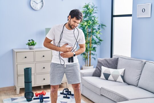 Handsome latin man wearing sportswear at home with hand on stomach because nausea, painful disease feeling unwell. ache concept.