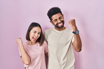 Young hispanic couple together over pink background very happy and excited doing winner gesture...