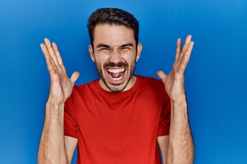 Naklejka premium Young hispanic man with beard wearing red t shirt over blue background celebrating mad and crazy for success with arms raised and closed eyes screaming excited. winner concept