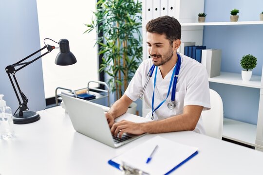 Young Hispanic Man Wearing Doctor Uniform Using Laptop Working At Clinic