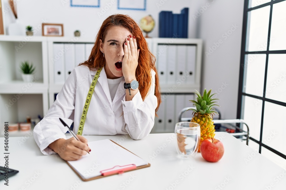 Wall mural Young redhead woman nutritionist doctor at the clinic yawning tired covering half face, eye and mouth with hand. face hurts in pain.