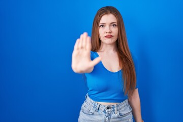 Redhead woman standing over blue background doing stop sing with palm of the hand. warning expression with negative and serious gesture on the face.