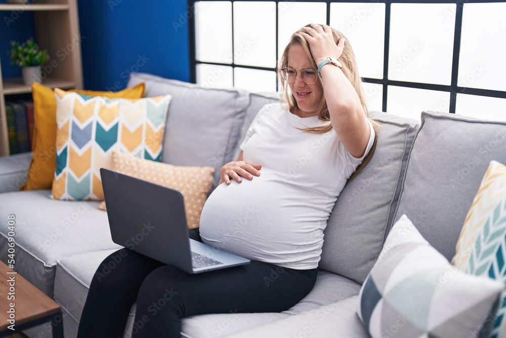 Wall mural young pregnant woman sitting on the sofa at home using laptop stressed and frustrated with hand on h