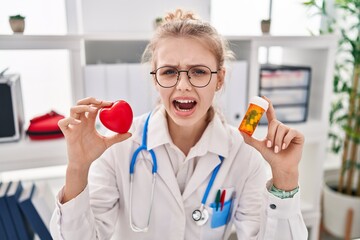 Young caucasian doctor woman holding pills and heart angry and mad screaming frustrated and furious, shouting with anger. rage and aggressive concept.