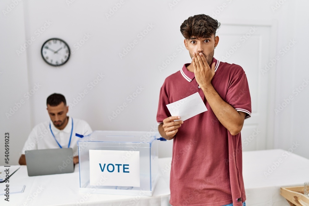 Sticker young hispanic man voting putting envelop in ballot box covering mouth with hand, shocked and afraid