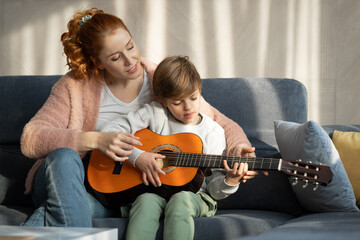 Happy mother teaching her son how to play the guitar in their living room on sunny day