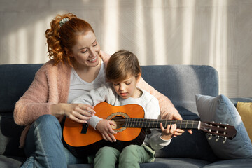 Happy mother teaching her son how to play the guitar in their living room on sunny day