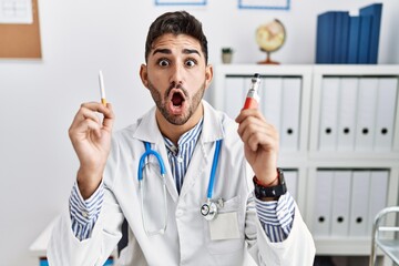 Young doctor man holding electronic cigarette at medical clinic afraid and shocked with surprise...
