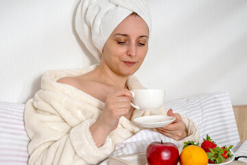 Obraz na płótnie Canvas Woman eating fruit breakfast in bed at home in the morning. Lady wrapping a towel around head. Girl wearing bathrobe on clean white bedding with cozy blanket. Healthy lifestyle