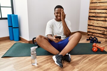 Young african man sitting on training mat at the gym using smartphone covering one eye with hand, confident smile on face and surprise emotion.