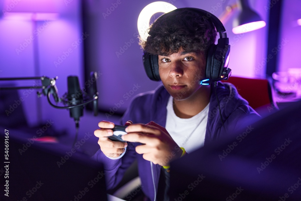 Wall mural young hispanic teenager streamer playing video game using joystick at gaming room