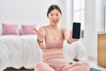 Chinese young woman holding smartphone showing blank screen pointing thumb up to the side smiling happy with open mouth