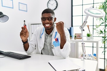 Young african american doctor man holding electronic cigarette at medical clinic screaming proud,...