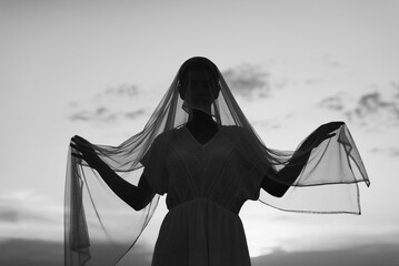 Silhouette of beautiful young woman in nature face covered with white silk veil against sky and clouds in black and white