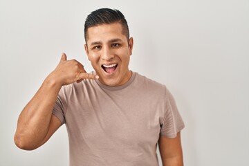 Hispanic young man standing over white background smiling doing phone gesture with hand and fingers like talking on the telephone. communicating concepts.