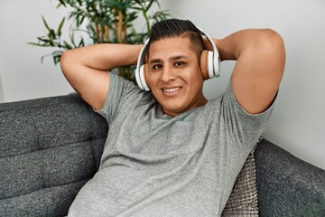 Young hispanic man smiling happy listening music sitting on the sofa at home.