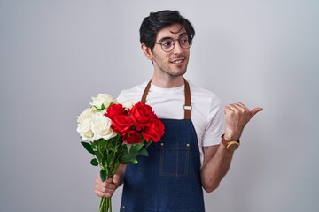 Young hispanic man holding bouquet of white and red roses smiling with happy face looking and pointing to the side with thumb up.