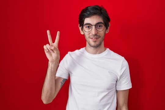 Young hispanic man standing over red background smiling looking to the camera showing fingers doing victory sign. number two.