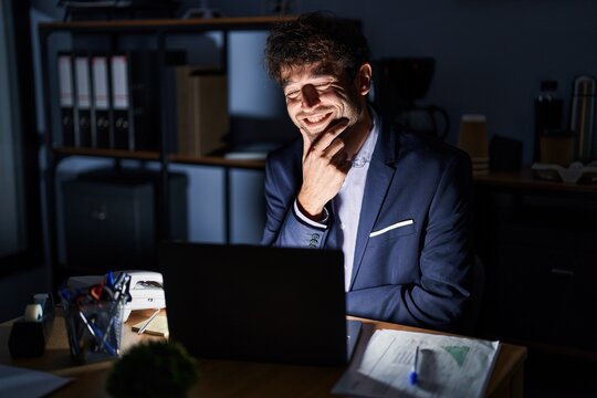 Hispanic young man working at the office at night looking confident at the camera smiling with crossed arms and hand raised on chin. thinking positive.