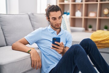 Obraz premium Young hispanic man using smartphone sitting on floor at home