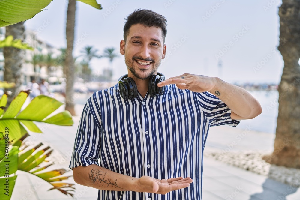 Sticker Young handsome man listening to music using headphones outdoors gesturing with hands showing big and large size sign, measure symbol. smiling looking at the camera. measuring concept.