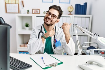 Fototapeta na wymiar Young man with beard wearing doctor uniform and stethoscope at the clinic pointing to the back behind with hand and thumbs up, smiling confident