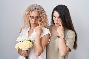 Mother and daughter holding bouquet of white flowers pointing to the eye watching you gesture, suspicious expression