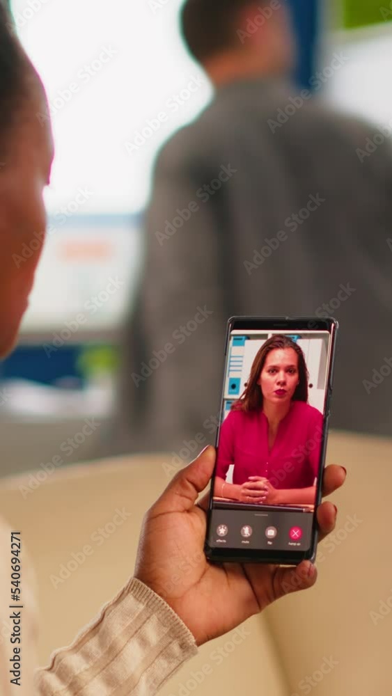 Poster back view of african woman discussing with remote manager on video call holding smartphone using hea