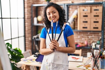 Young chinese woman artist smiling confident holding paintbrushes at art studio