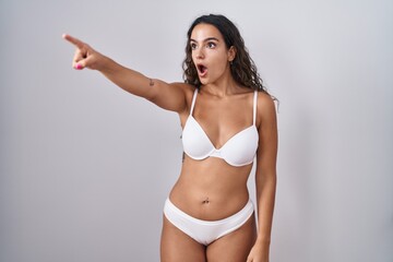 Fototapeta premium Young hispanic woman wearing white lingerie pointing with finger surprised ahead, open mouth amazed expression, something on the front