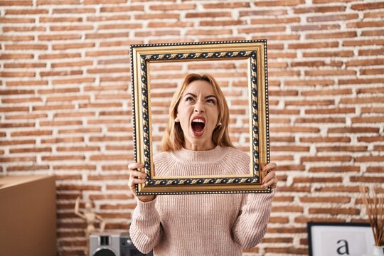 Hispanic woman holding empty frame angry and mad screaming frustrated and furious, shouting with anger looking up.