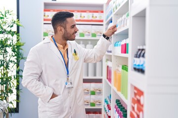 Young hispanic man pharmacist smiling confident holding product on shelving at pharmacy