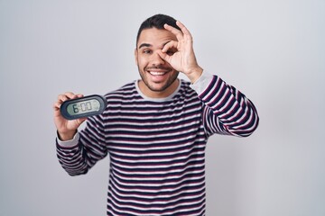 Fototapeta premium Young hispanic man wearing pyjama holding alarm clock smiling happy doing ok sign with hand on eye looking through fingers
