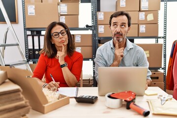Middle age couple working at small business ecommerce thinking concentrated about doubt with finger...